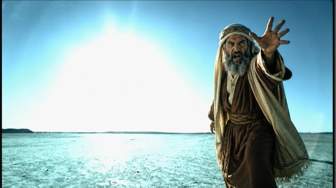 A bearded man in traditional Middle Eastern robes stands dramatically in a vast, sun-drenched desert, reaching one hand toward the camera with an intense, commanding expression. The image evokes the biblical story of Moses, visually reinforcing the message "NEVER BE AFRAID TO USE YOUR NAME" by depicting a prophet boldly declaring his identity and authority.