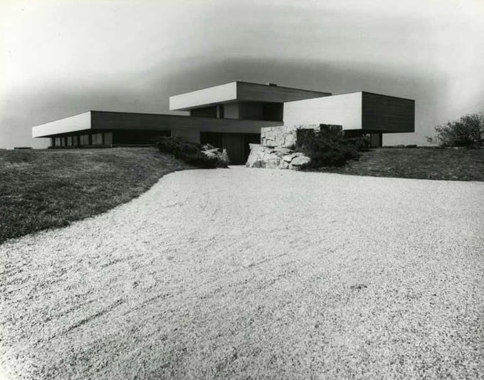 A black-and-white vintage photograph of a modernist house with flat, cantilevered roofs perched on a hill, approached by a curved gravel driveway.