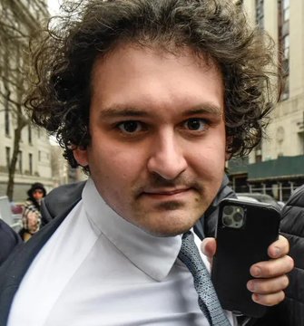 A close-up photo of a man with curly hair wearing a suit and tie, holding a black smartphone on a city street.
