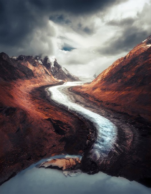 A dramatic landscape painting or digital artwork depicts a winding icy river or glacier cutting through rust-red mountains under heavy, luminous clouds.