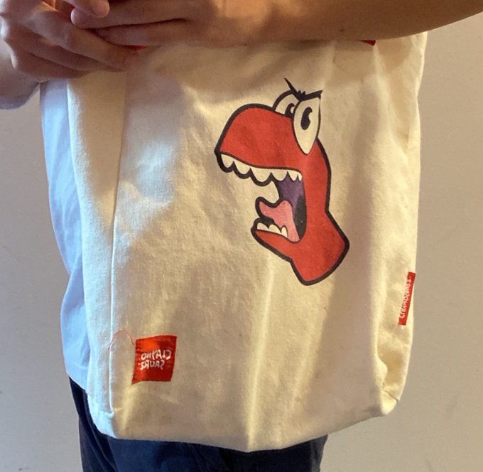 A person holding up a cream-colored tote bag featuring a cartoon illustration of a red dinosaur or creature with an open mouth, with red brand labels visible.