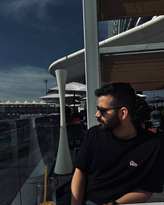 A bearded man in sunglasses and a black t-shirt sitting at an outdoor café with a glass and straw, overlooking a modern architectural structure under a blue sky.