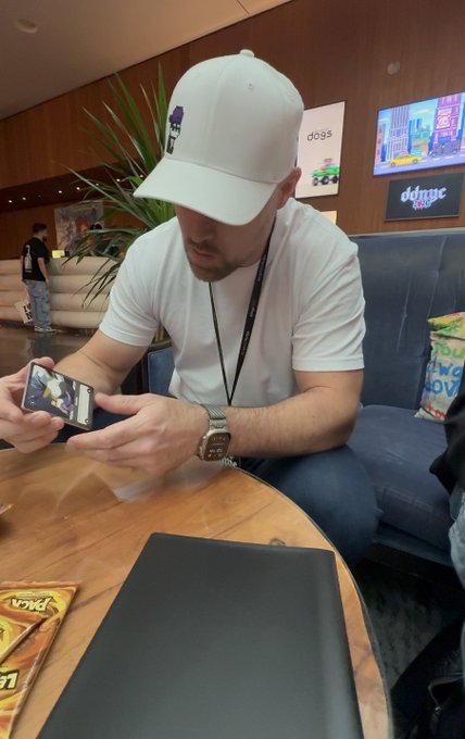 A man wearing a white baseball cap and a white t-shirt sits in a blue lounge chair at an event, looking down at his smartphone, with a coffee table and snack bags in the foreground and digital displays reading 'dogs' in the background.