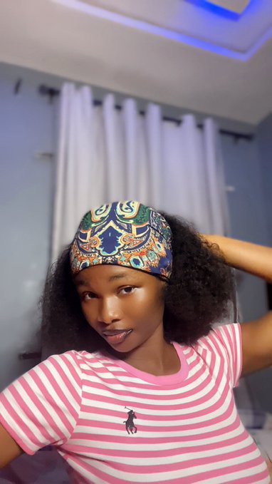 A woman wearing a colorful patterned headband and a pink and white striped Polo Ralph Lauren t-shirt poses indoors against a backdrop of white curtains.