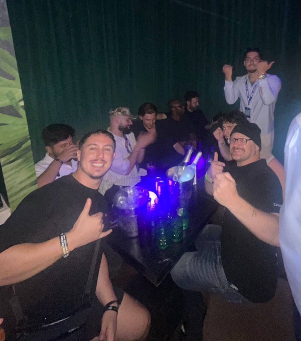 A group of people pose together at a table with bottles and blue-lit glasses, smiling and making hand gestures in a dark setting with green curtains.