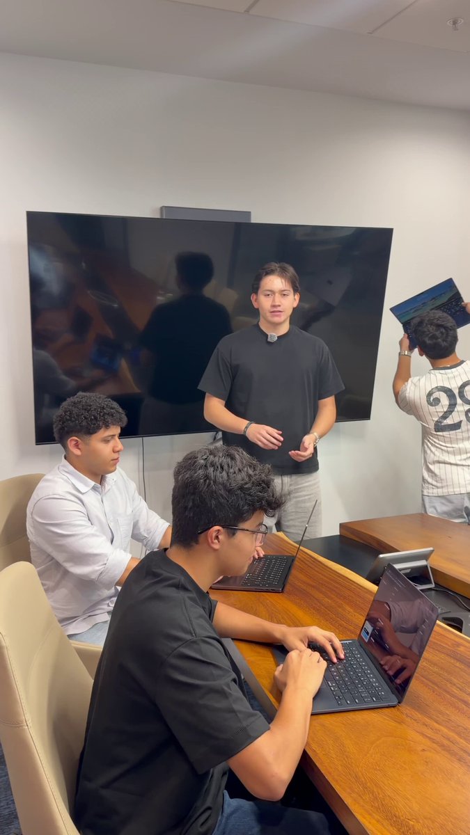 The image shows a group of people sitting around a table, with one person using a laptop computer while others are engaged in conversation. The scene appears to be taking place in an office setting or conference room, as there is a large screen TV mounted on the wall behind them. The people seem to be discussing something related to finance and real-world assets, possibly talking about Neko Proteo, a Costa Rican team building company that offers on-chain yield generation services without requiring users to understand DeFi.