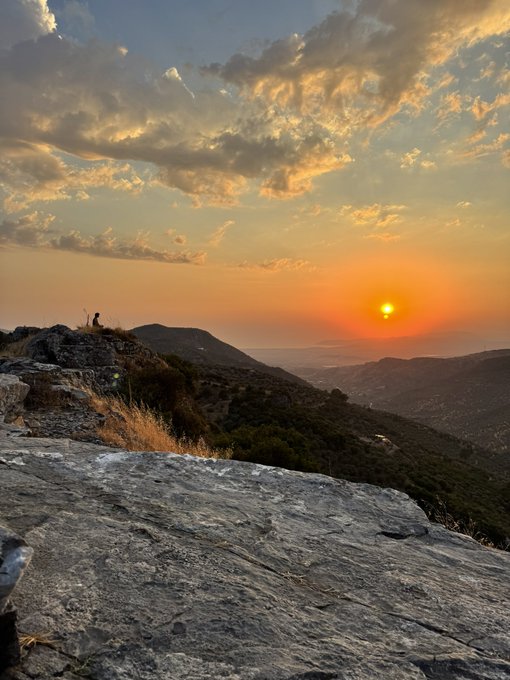 A scenic photograph of a sunset over a rocky mountain landscape, with the sun glowing on the horizon beneath dramatic clouds.