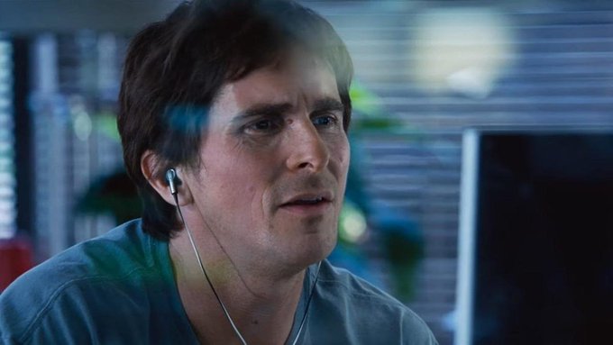 A man with brown hair and a mustache, wearing a blue shirt and white wired earbuds, looks intently off-camera in what appears to be an office setting.