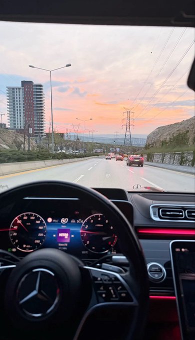 A view from inside a Mercedes car driving on a highway at sunset, showing the dashboard and steering wheel with the city skyline and power lines visible ahead.