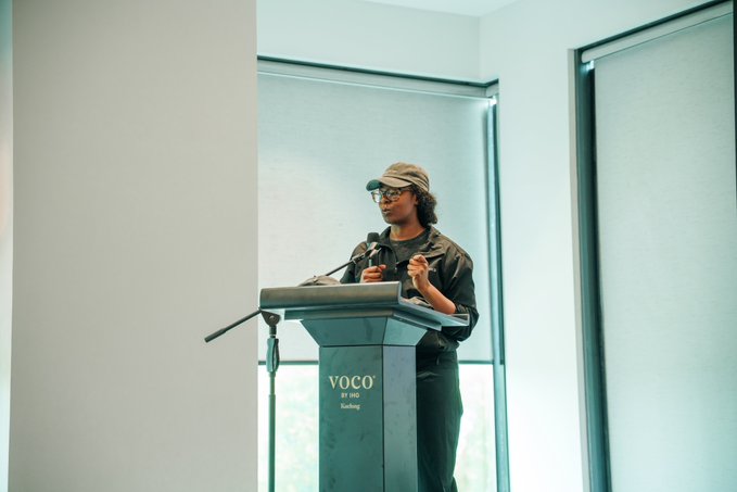 A woman wearing a cap and glasses speaking at a podium labeled 'VOCO BY IHG Darling', gesturing with her hand during what appears to be a presentation.