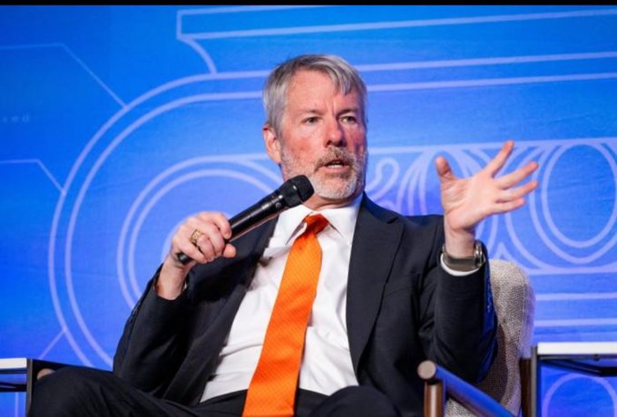 A gray-bearded man in a dark suit and bright orange tie speaks into a microphone, gesturing with his hand while seated on stage against a blue backdrop featuring geometric designs. The image relates to the post by depicting what appears to be Michael Saylor, the executive behind Strategy's massive Bitcoin accumulation highlighted in the accompanying text.