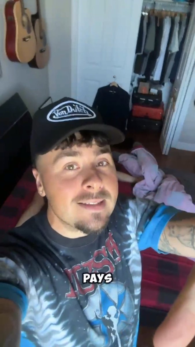 A young man wearing a Von Dutch cap and a graphic t-shirt takes a selfie in his bedroom, with guitars mounted on the wall behind him and an open closet visible. The word "PAYS" appears in bold white text at the bottom, tying into his post about giving $100 to a stranger and promoting his rugs.fun link.