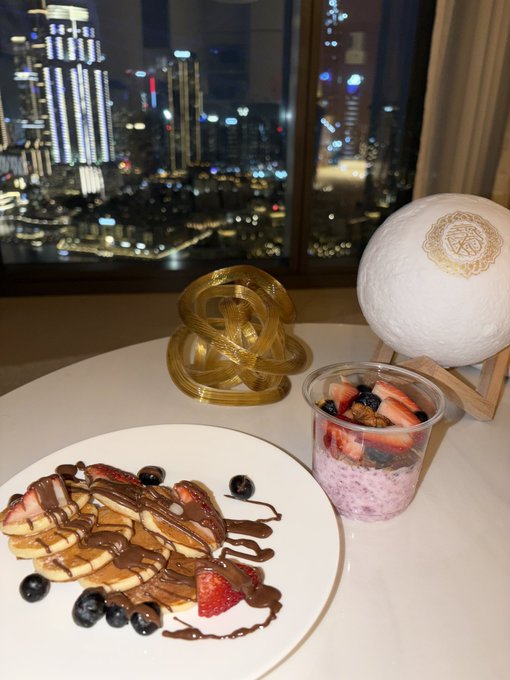 A stack of chocolate-drizzled pancakes with strawberries and blueberries sits on a white plate alongside a berry parfait cup, a gold decorative knot, and a glowing moon lamp, all set against a stunning nighttime city skyline view. The luxurious room service breakfast mirrors the celebratory tone of the post about $SOL rebounding to $280, suggesting an indulgent treat to mark the crypto milestone.