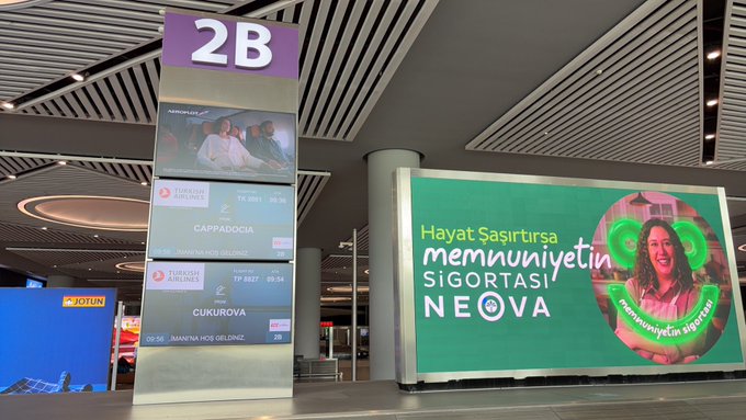 The image shows a large green sign with white text that says "Hayat Sigurtirasımünüyoruz." The sign is located inside an airport terminal, possibly near the baggage claim area or waiting room. A person's face can be seen in the background of the image, but it does not provide enough information to determine their role within the scene.