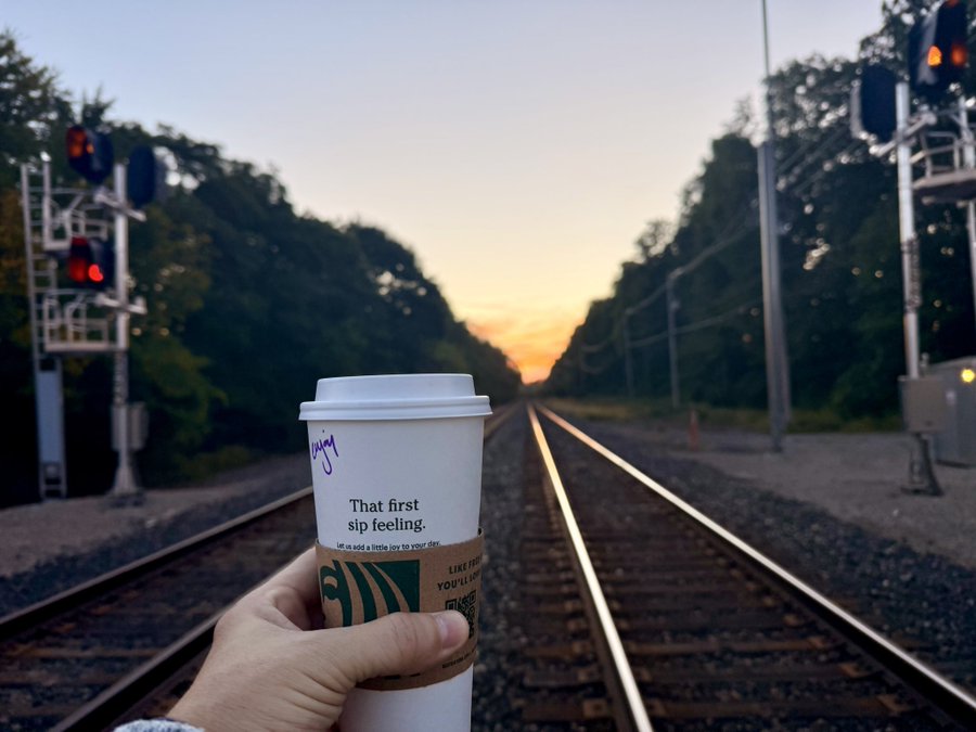 A hand holds a white Starbucks cup reading 'That first sip feeling. Let us add a little joy to your day.' in front of railway tracks at sunset.