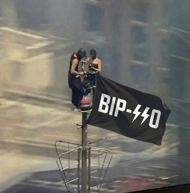 A photograph of two people perched atop a street light pole holding a black flag that reads 'BIP-440,' referencing a Bitcoin Improvement Proposal.