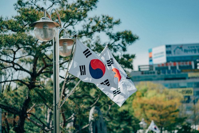Two South Korean national flags wave from a lamppost against a backdrop of trees and a city building, visually grounding a story about South Korea's Supreme Court proposing formal rules for seizing and liquidating crypto holdings, with a public comment period closing August 11.
