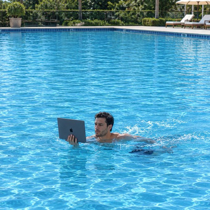 A man floats in a bright blue swimming pool while holding and looking at an open Apple laptop, with lounge chairs and umbrellas visible in the background.