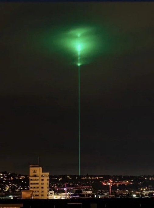 A night photograph of a city skyline where a bright, glowing green laser beam shoots vertically from the ground into the dark sky, creating a distinct illuminated spot high above the urban landscape.