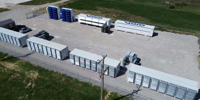 An aerial view of an industrial storage facility featuring rows of white shipping containers, two large white trailers labeled 'GIGA', and stacks of blue and yellow barrels on a gravel lot.