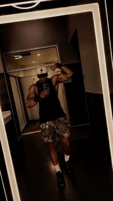 A muscular man takes a mirror selfie while flexing his bicep, wearing a black tank top, camo shorts, a cap, and white socks in what appears to be a modern bathroom or locker room.