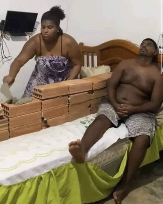 A woman stands next to a bed built from stacked bricks and a mattress, where a shirtless man is lying down with a distressed expression.
