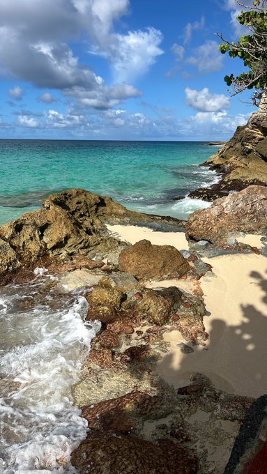 A scenic coastal view featuring turquoise waters, white-capped waves crashing against rugged rocky outcrops and a small sandy beach under a blue sky with scattered clouds.