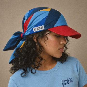 A person with curly dark hair wearing a blue, red, and yellow striped headscarf/bandana and a light blue t-shirt, looking down with their eyes closed.
