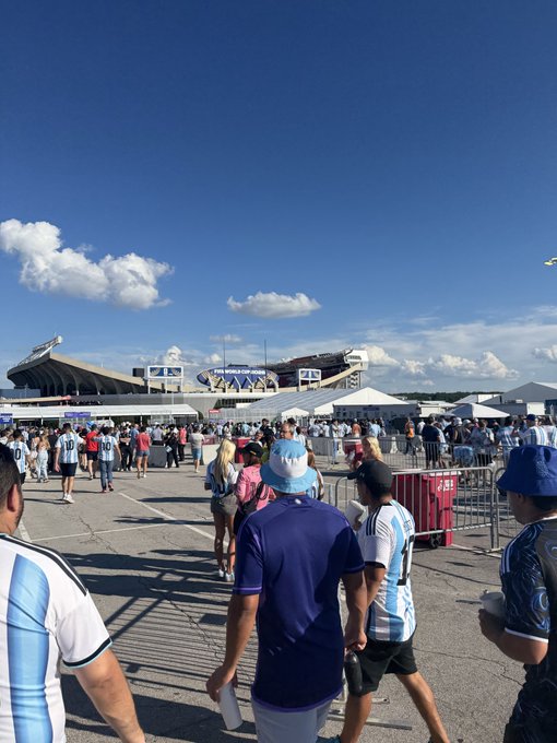 Crowds of NASCAR fans, many wearing blue and white gear, walk toward a racetrack stadium under a partly cloudy sky. The energetic atmosphere and racing venue provide an ironic backdrop, emphasizing the post's point that the crowd remains unaware of the substantial $200M valuation of $CASHCAT hidden beneath the mainstream spectacle.