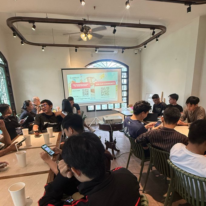 A group of young people seated around tables in a cafe or co-working space, watching a presentation displayed on a projector screen with QR codes labeled 'JOIN OUR COMMUNITY'.