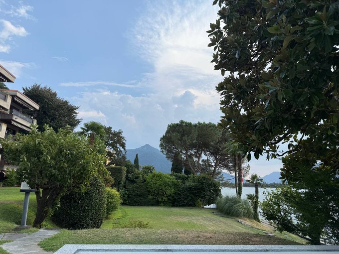 A serene lakeside garden scene features lush green hedges, manicured lawns, and tall trees framing a view of distant mountains under a partly cloudy sky, with a building partially visible on the left. The tranquil, picturesque setting perfectly matches the post's invitation to enjoy a beautiful day in such an idyllic location.