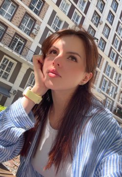 A young woman with long brown hair poses for a selfie on a sunny day, wearing a blue and white striped shirt and a yellow watch. The cheerful outdoor selfie complements the upbeat "Happy #TEZOSTUESDAY" message, capturing a bright and positive mood to start the day.