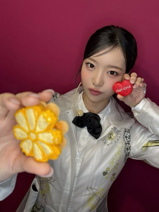 A young woman with dark hair poses against a magenta background holding a sliced orange in one hand and a red heart-shaped object reading 'HIGH-LOVE' near her face, wearing a white patterned jacket.