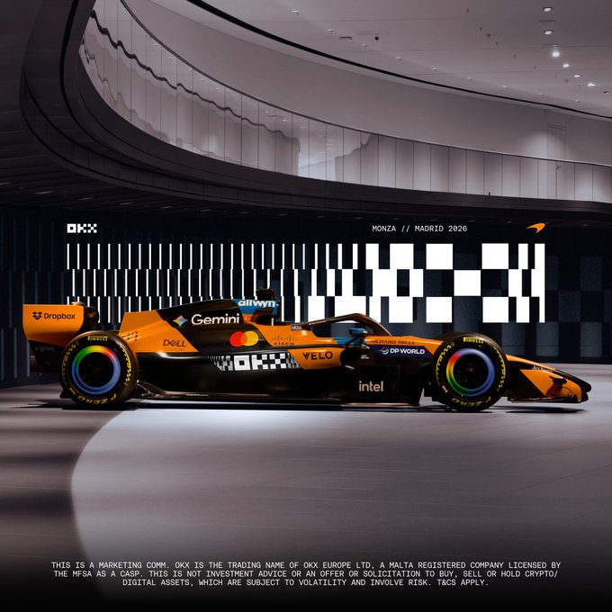 A McLaren Formula 1 car in a papaya orange and black livery displayed in a modern showroom setting, branded with sponsors including Dropbox, Dell, Gemini, Gillette, OKX, Yelo, and Intel, with 'MONZA // MADRID 2026' on the wall.