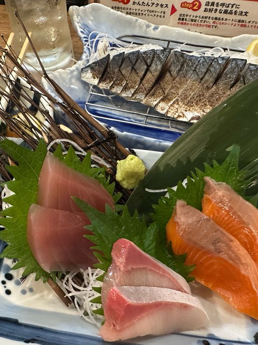 A Japanese sashimi platter featuring sliced raw fish (tuna, salmon, and yellowtail) on shiso leaves, with wasabi and grilled fish in the background, along with access instructions in Japanese.