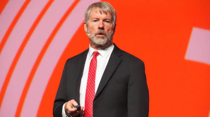 A man with gray hair and beard, wearing a black suit, white shirt, and red tie, speaks on a stage with an orange background while holding a clicker. The image likely depicts Michael Saylor, the Executive Chairman of MicroStrategy, referenced in the post about using ChatGPT to design preferred shares like $STRC for raising capital to buy more bitcoin.