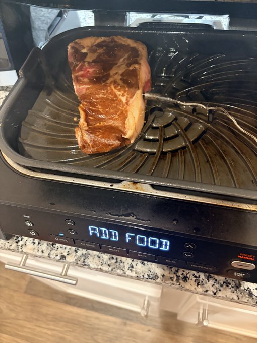 A cooked steak with a meat thermometer probe sits in an air fryer with 'ADD FOOD' displayed on its digital screen.