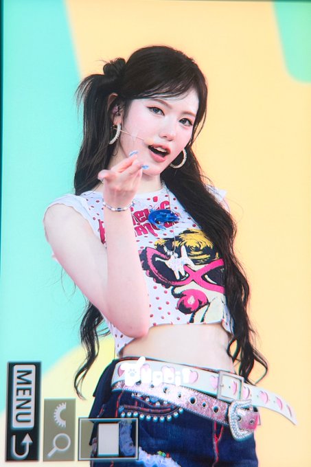 The image shows a performer with long dark hair in pigtails wearing a colorful crop top with polka dots and graphic prints, along with a studded belt. The interface displays "MENU" and "260731" as visible text, suggesting this is a screenshot from a Tokyo Idol Festival (TIF) broadcast. The caption references SUI from KiiiKiii performing at the event, consistent with the on-stage idol performance captured in the frame.