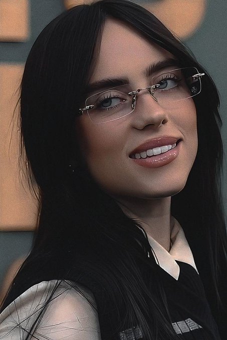 A close-up portrait of a dark-haired woman wearing rimless glasses and a black vest over a white shirt, smiling slightly.