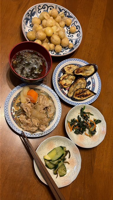 An overhead view of a traditional Japanese home-cooked meal featuring simmered potatoes, miso soup, grilled eggplant, stewed meat with vegetables, pickled spinach with carrots, and pickled cucumber on a wooden table.