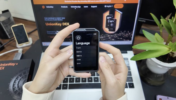 A person holds a UnionKey DEX hardware wallet displaying a language selection menu (English, 简体中文, 繁體中文, 日本語, 한국어) in front of a laptop showing the UnionKey DEX website, which the caption suggests is nearly identical to OneKey's open-source hardware and firmware.