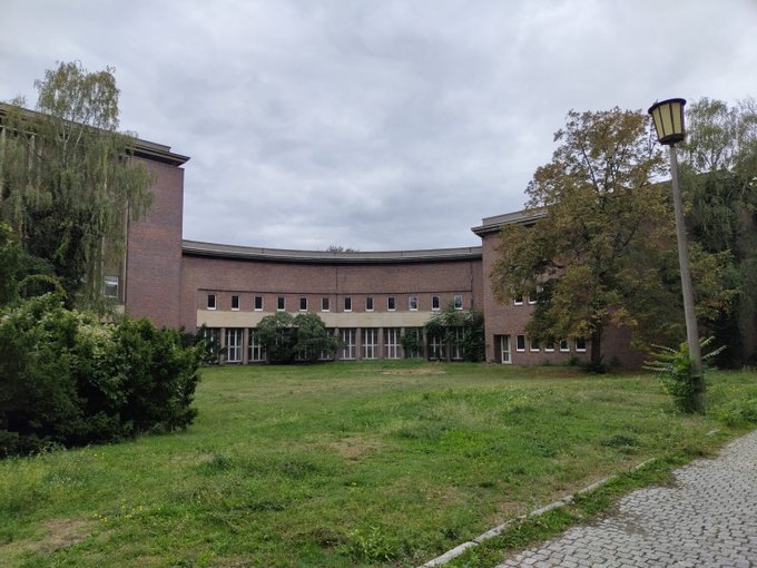 An exterior view of a curved brick building with multiple windows, surrounded by trees, bushes, and a lawn under a cloudy sky, with a street lamp on the right.