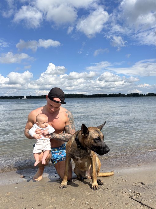 A shirtless man in a cap and swim trunks kneels on a sandy beach holding a baby in white, with a large brown dog sitting beside them against a backdrop of water, distant sailboats, and a partly cloudy sky—capturing the relaxed "beach day with the boys" vibe described in the post, where "the boys" appear to be his infant son and his loyal canine companion enjoying crypto gains and sunshine.