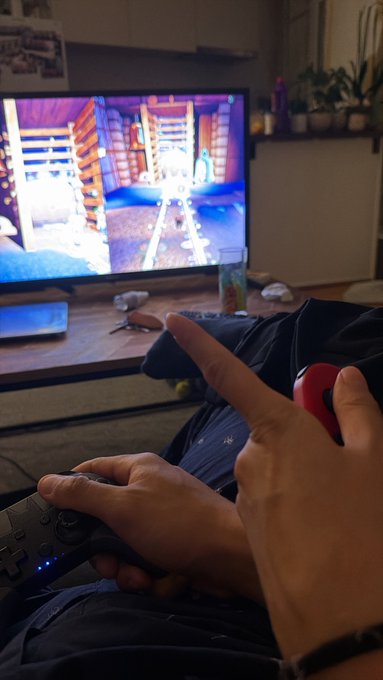 A person holds a Nintendo Switch controller in one hand while pointing a finger at the screen, which shows a brightly colored video game.