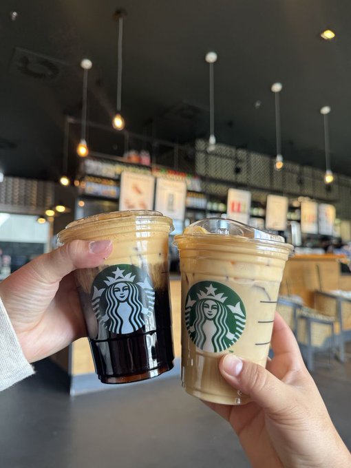 Two hands clink clear plastic Starbucks cups—one containing dark cold brew and the other an iced latte—in a toast inside a modern café. The Starbucks siren logo is prominently displayed on both cups, while the caption humorously contrasts the casual coffee meetup with the inevitable deep-dive conversations about crypto and AI.