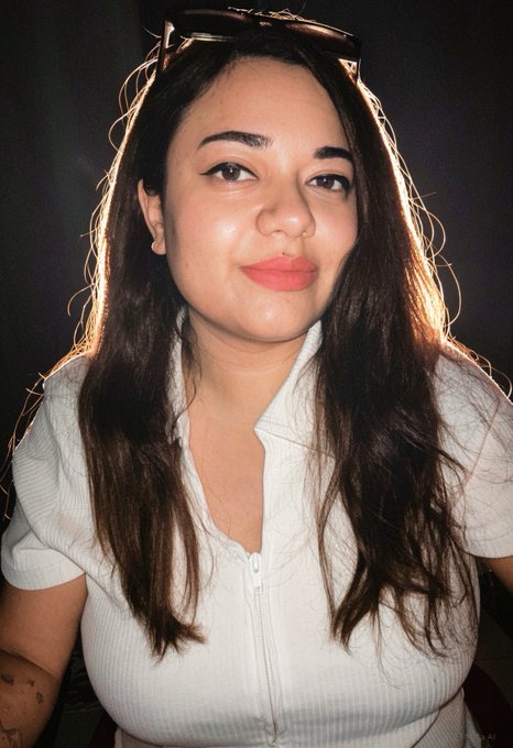 A selfie of a smiling young woman with long dark hair, sunglasses perched on her head, and red lipstick, wearing a white ribbed zip-up top.
