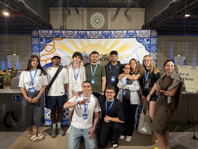 A group of around ten young people wearing lanyards pose together indoors in front of a decorative blue and yellow mural that reads 'Global [Lis]bon'.