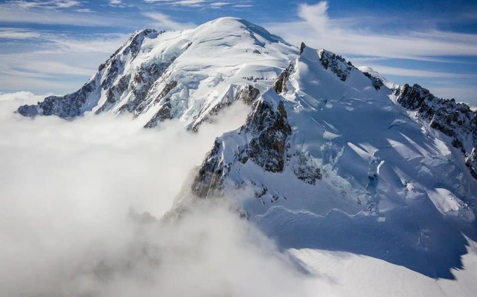 A stunning aerial view of a massive snow-capped mountain peak rising above a thick layer of clouds under a blue sky with scattered white clouds.