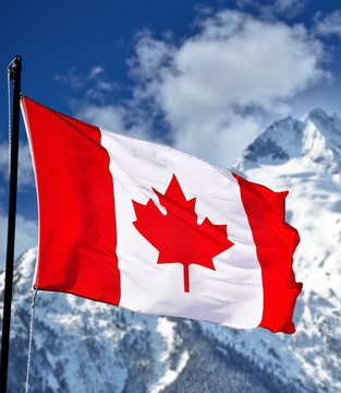 The Canadian flag waving against a backdrop of snow-capped mountains and blue sky with white clouds.