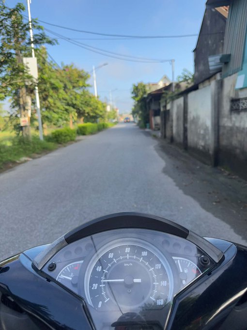 A first-person view from a motorbike showing the speedometer and dashboard in the foreground, looking down a quiet rural road lined with trees and houses.